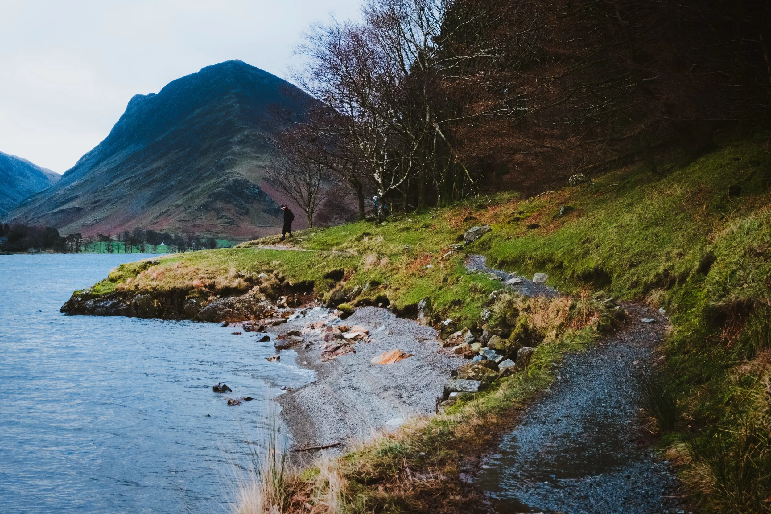 Two older hikers make their way to the shore of Buttermere with Fleetwith Pike in the distance.