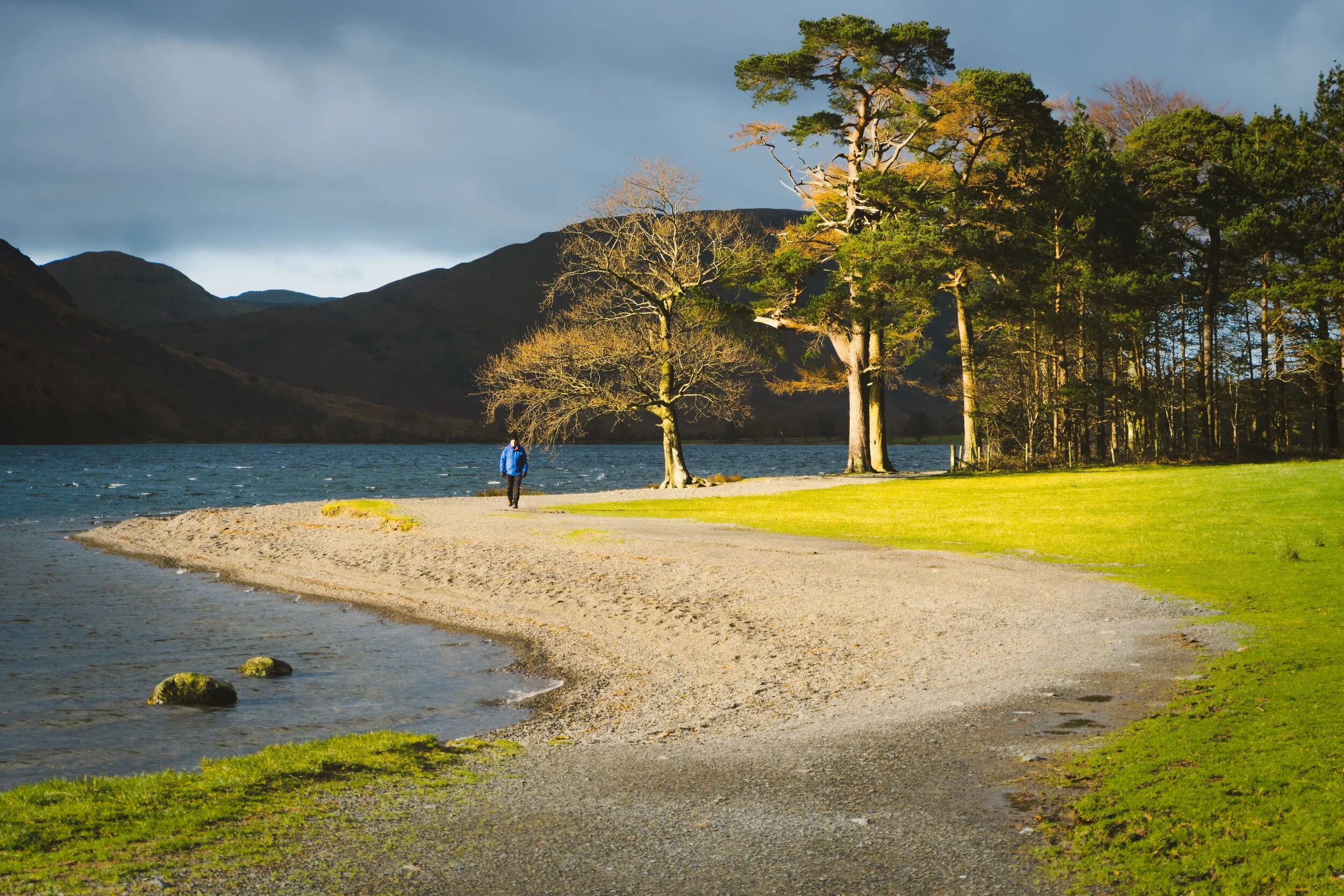 Looking south from the head of Buttermere, a burst of light illuminates some trees and a hiker near the shore.