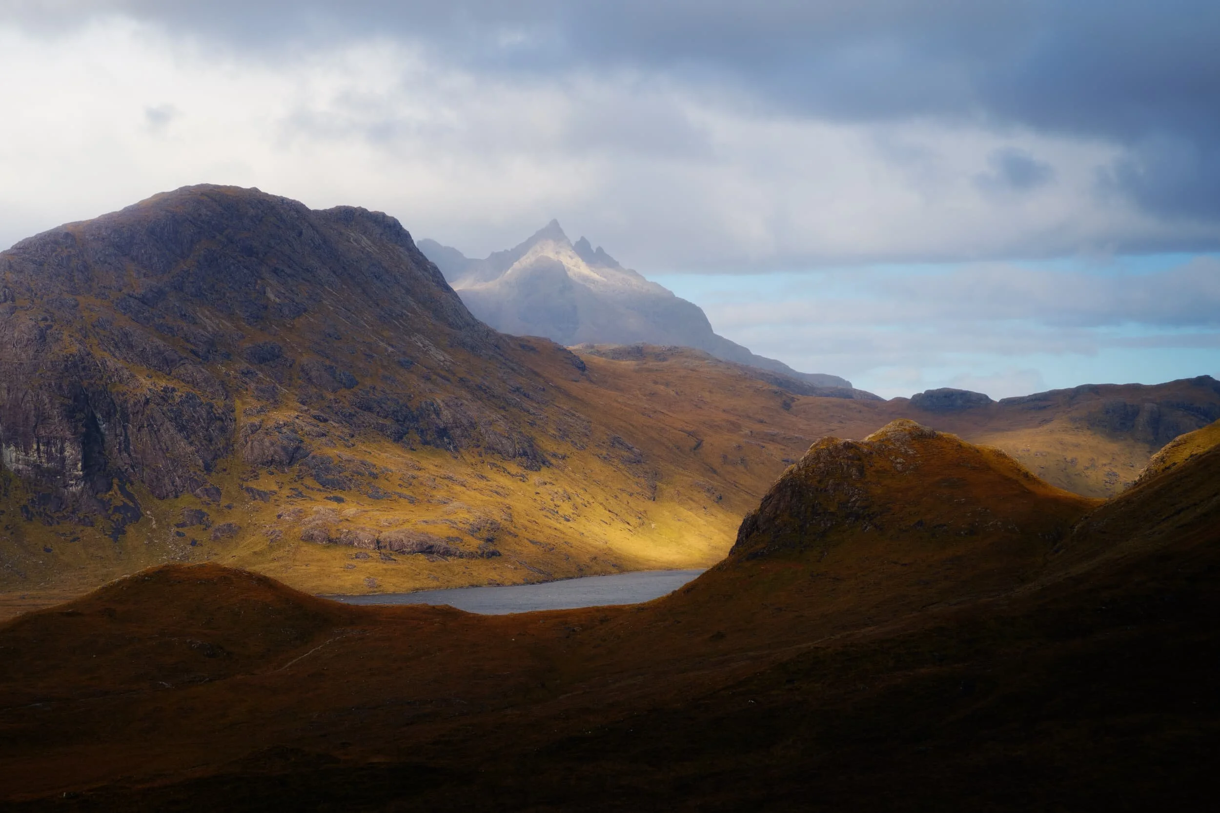  A particularly strong burst of light breaks through the low clouds as I shot this view across  Loch na Creìtheach . The hill on the left is  Sgùrr Hain  (420 m/1,377 ft) and the crown-like summit in the distance is  Sgùrr nan Gillean  (966 m/3,170 ft), one of Skye&rsquo;s most famous peaks. Its name translates from Scottish Gaelic as &ldquo;peak of the young men&rdquo;. 