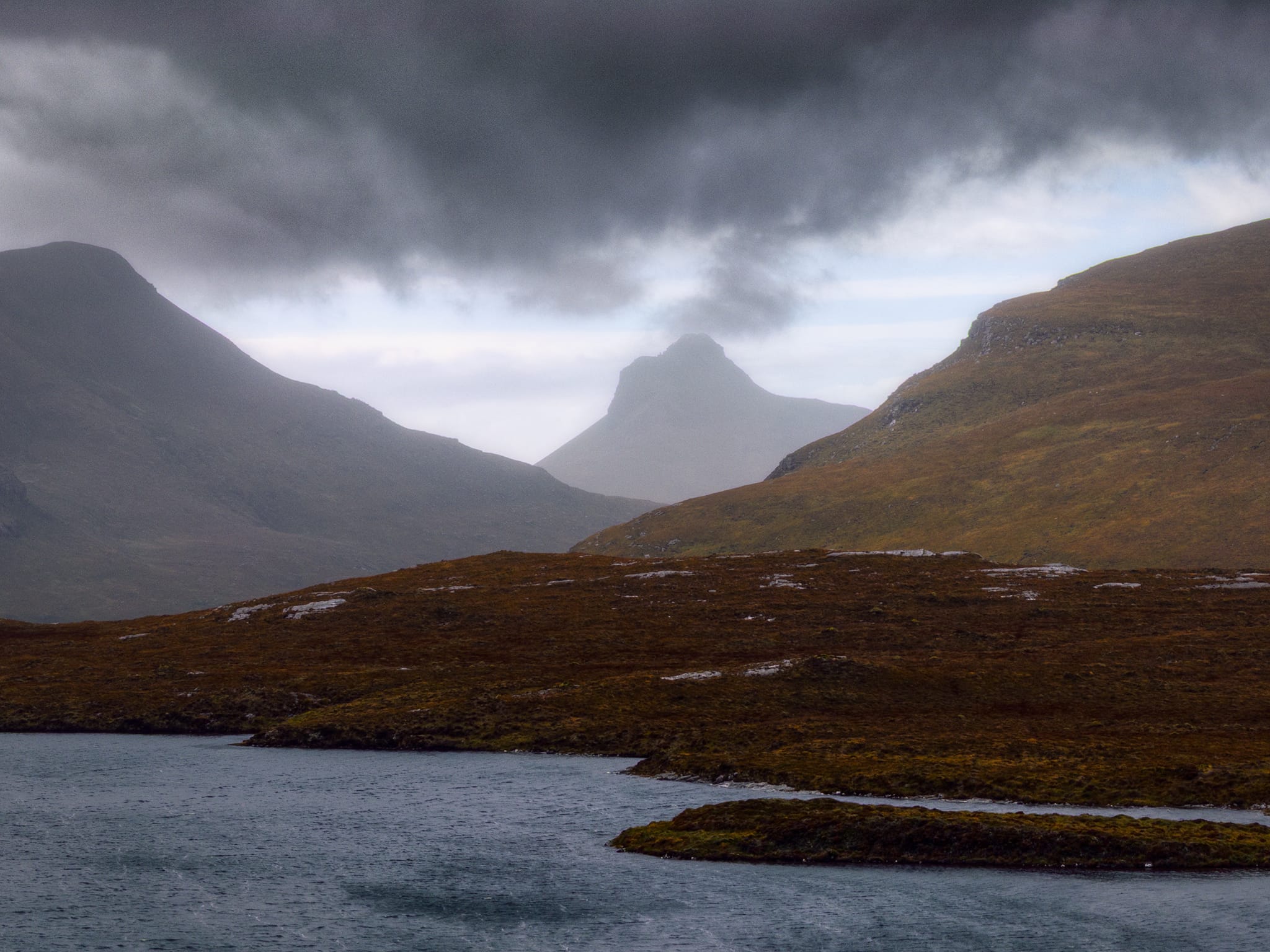At the Knockan Crag Nature Reserve we take in the glorious sight across Lochan an Ais of Stac Pollaidh (612.4 m/2,009 ft), framed between the shoulders of Cùl Beag, (769 m/2,523 ft) and Cùl Mòr (849 m/2,785 ft). The driving storm clouds give Stac Pollaidh the impression that it’s a volcano spewing ash into the sky.