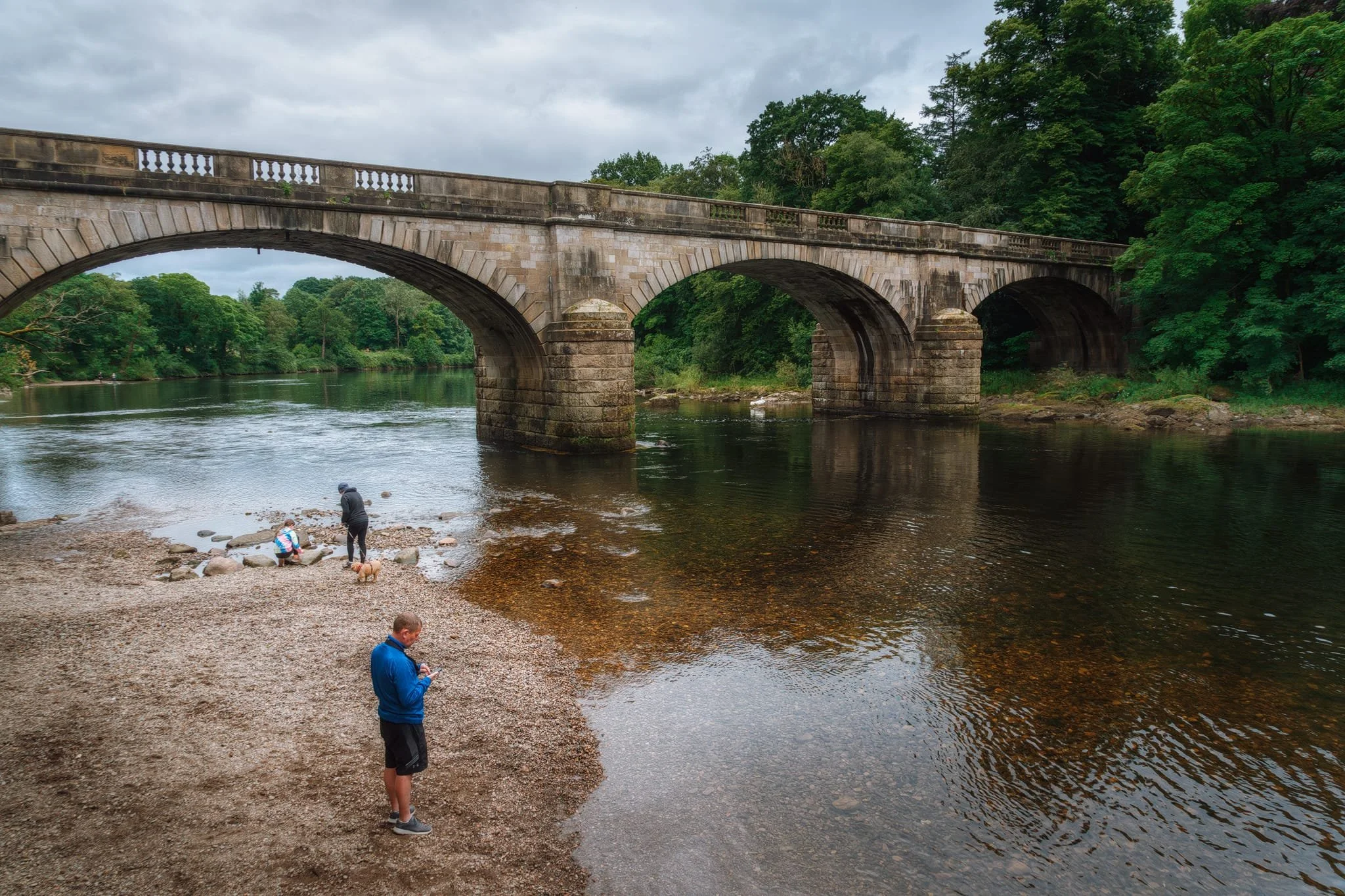  Retracing our steps, we crossed back over the old railway viaduct to locate the footpath alongside the river. At the river shore, families and dogs played around in the water with the Caton Lune Bridge standing above. This is a Grade II listed road bridge. The original bridge at this spot was built privately in 1806. It was a stone structure with three arches, nicknamed 