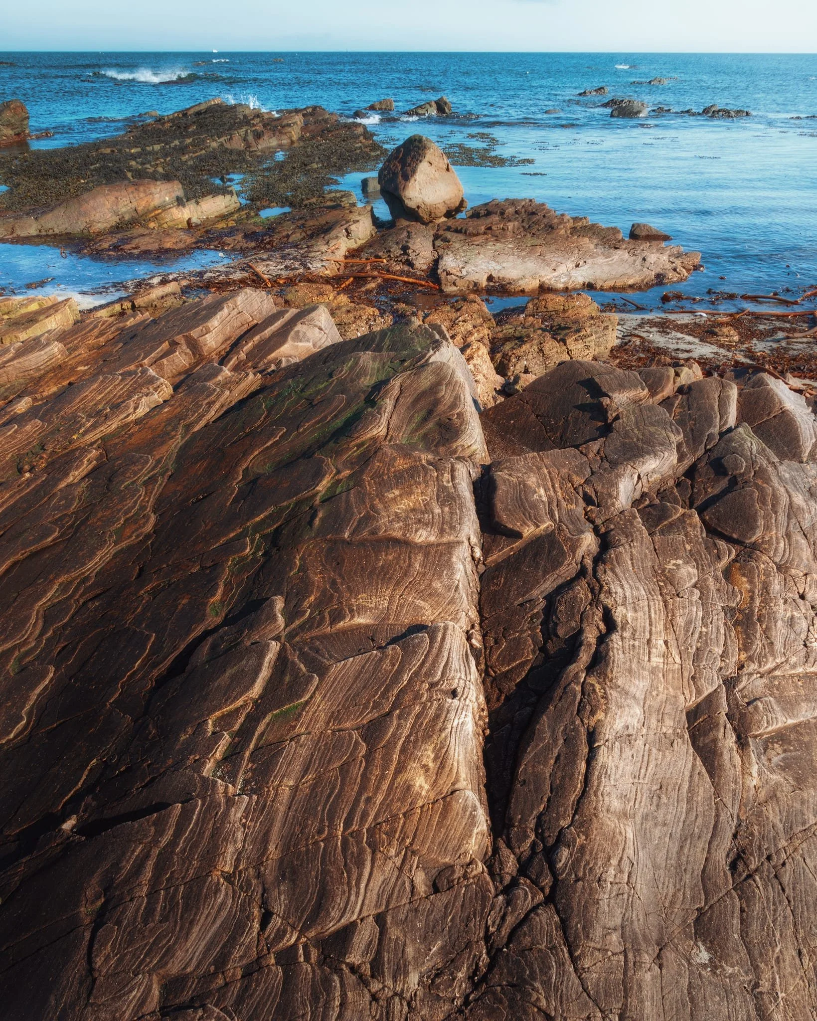 Striking rock formations with wavy patterns and noticeable cracks. Close up you can see twisted layers of metamorphic rock that look like thin layered sheets. These layers show signs of ancient geological activity. The swirling shapes are created by uneven erosion, which points to different minerals in the rocks. Meanwhile, the beautiful blue waters around continue to shape these rocks through their ongoing movement and waves.