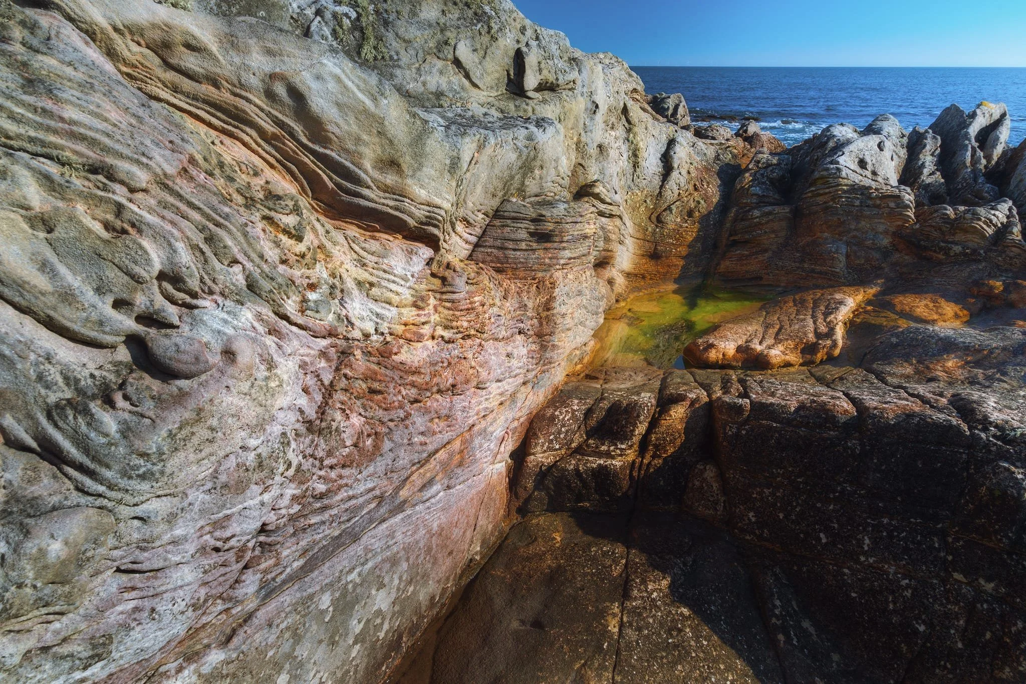 At Lochaber Rock, beautiful layers can be seen in the light-colored sandstone, with reddish-pink stripes. On the left side, you can see honeycomb-like patterns and wavy textures, which look different from the darker, sharp shapes on the right. These differences show how the rocks are made of different materials and react in unique ways to the ocean’s wear.