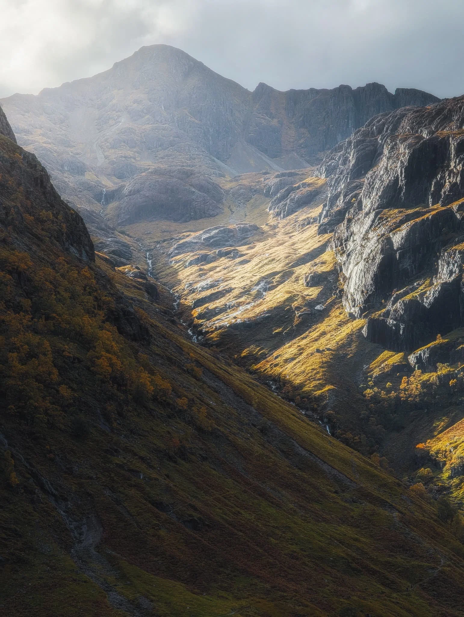  In the heart of Glencoe we stopped at the main car park and quickly located the trail east towards the head of the valley. Looming above us were the Three Sisters of Glencoe. With light piercing through the clouds above, I captured this 5x photo of  Corrie nan Lochan&rsquo;s  crags getting drenched in light with  Stob Corrie nan Lochan  (1,115 m/3,658 ft) rising above. 