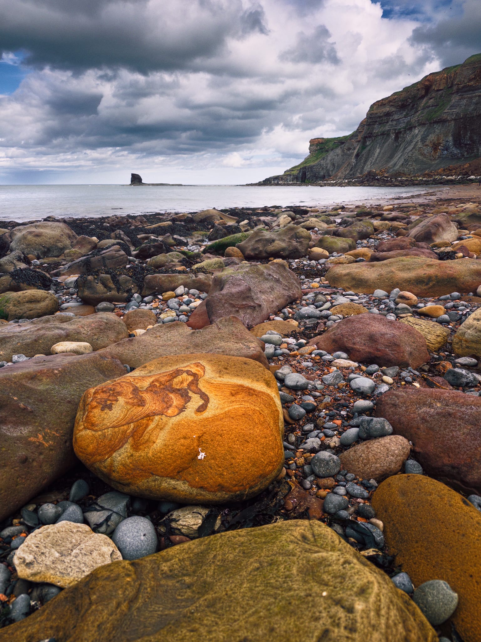 The bay is littered with beautifully sculpted rocks and boulders, including this lovely golden sandstone boulder. The swirling orange/brown pattern is Liesegang banding, rings of iron oxide (limonite/goethite) precipitated by groundwater diffusing through the porous rock after it was already cemented. In the distance, Black Nab all on its own.