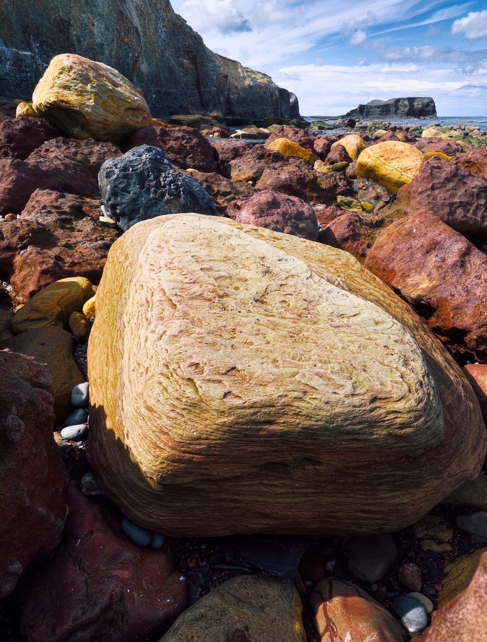Now making my way to the northeastern cliffs, gingerly picking my way through the boulder field and finding compositions towards Saltwick Bay. Here the same iron-oxide chemistry gives some lovely colours: red/purple boulders are iron-rich sandstone, buff/gold ones are more leached, the swirled foreground boulder shows Liesegang banding, and the dark grey boulder is likely shale.