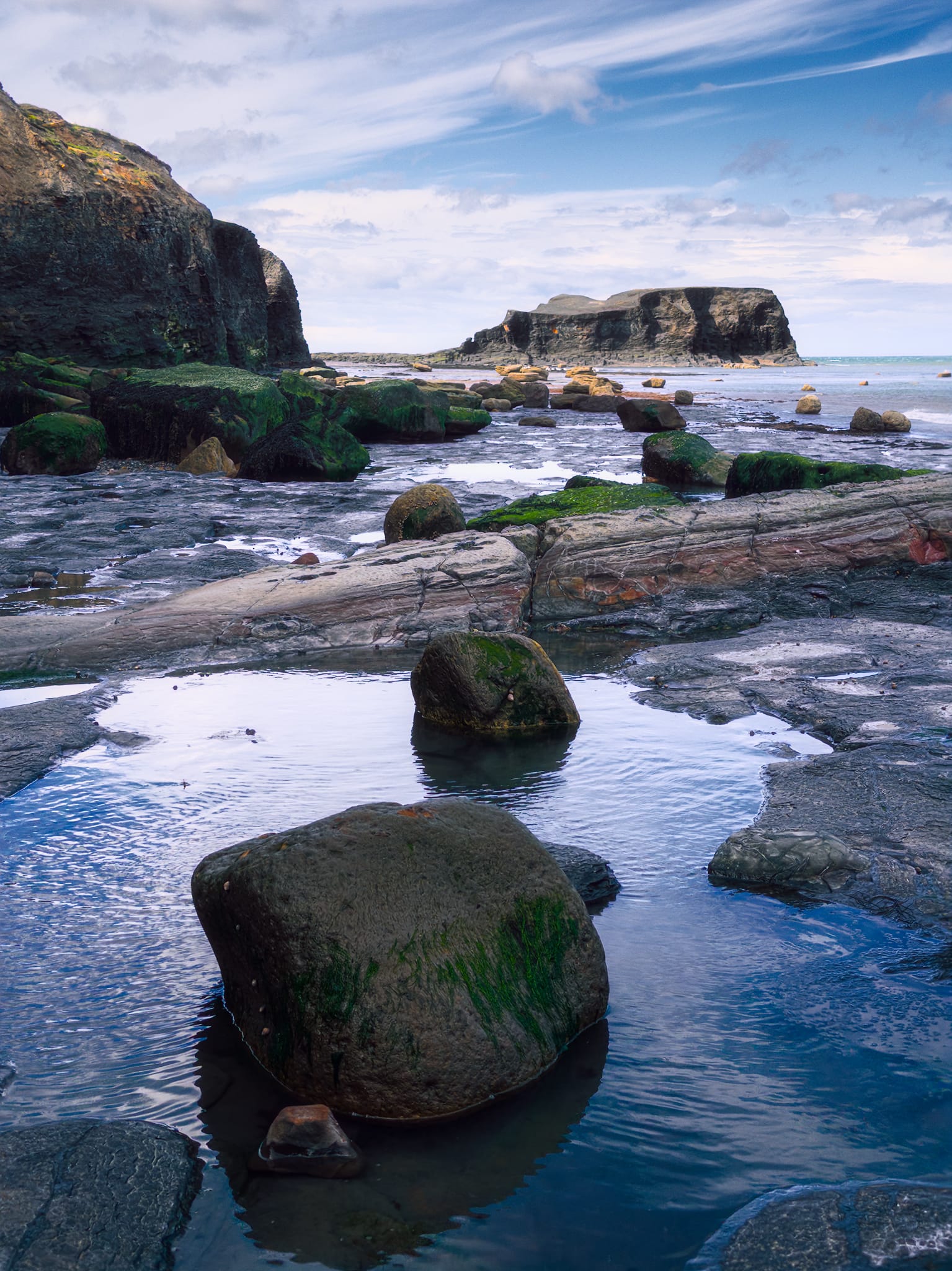 That&rsquo;s Saltwick Nab in the distance, the flat-topped headland (&ldquo;North Batts&rdquo; area). A beautiful wave-cut shale platform in the foreground cuts across with algae-covered boulders sitting in tidal pools. Green algae marks tidal exposure zones; the smooth, flat bedding surfaces are Alum Shale, kept polished by regular wave action.