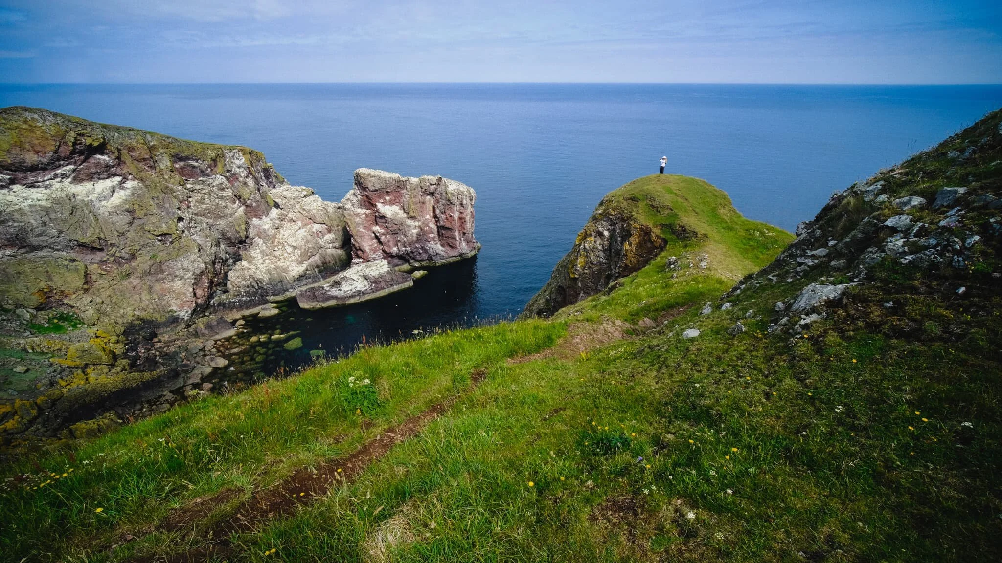  Beyond the lighthouse, the cliffs get steeper and more vertical. Lisabet follows one spur alone. Click/tap the image for a bigger view; you may well be able to point out the hundreds of birds on the cliffs to the left. 