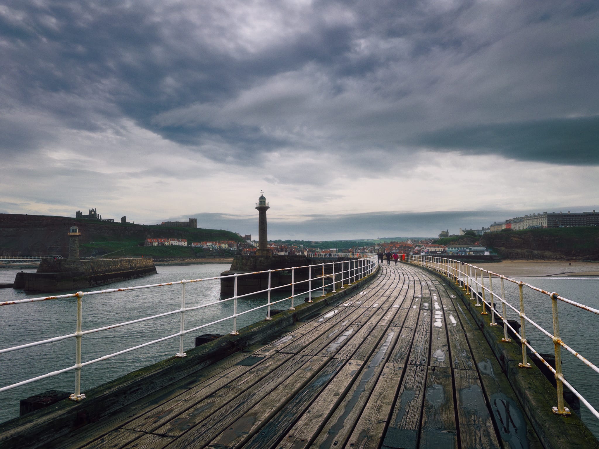 Looking back from the end of the West Pier, clouds racing above. On the cliffs to the far left is Whitby Abbey with St Mary&rsquo;s Church a little further in. To the left in the sea is Whitby East Pier Lighthouse, with the West Pier Lighthouse more central. To the right on the cliff is the newer part of Whitby, with Victorian buildings constructed to accommodate Whitby&rsquo;s then growing status as a tourist destination.